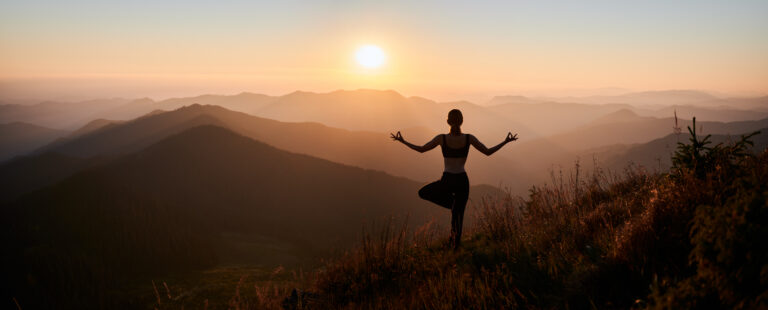 Eine Frau meditiert in der Natur auf einem Bein stehend.